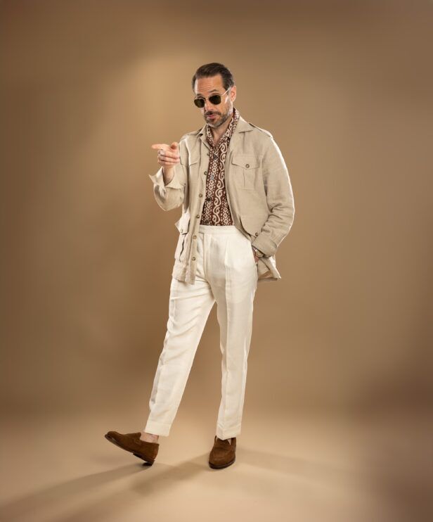 Beige linen safari jacket with brown printed shirt, cream pleated trousers and tan suede loafers, worn by a model