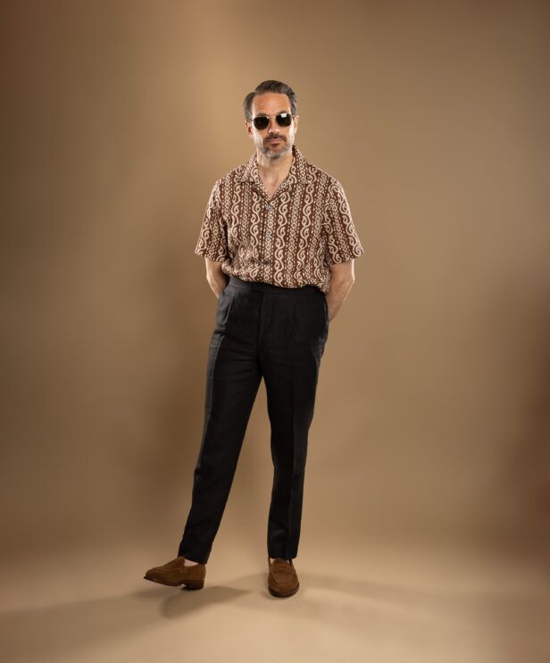 Brown and cream printed camp collar short sleeve shirt with black linen trousers and tan suede loafers, worn by a model