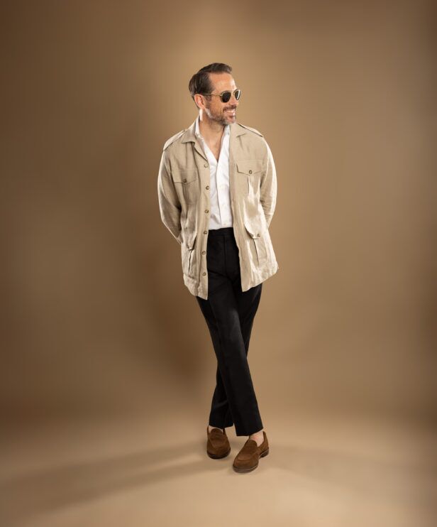 Beige linen safari jacket with white shirt, black trousers and brown suede loafers, worn by a model