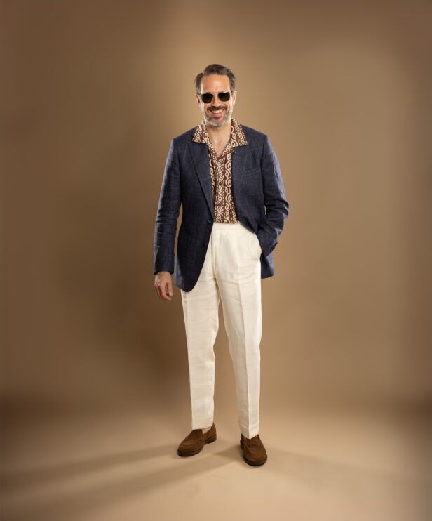 Navy checked linen sport coat with brown patterned camp shirt, cream trousers and suede loafers, worn by a model