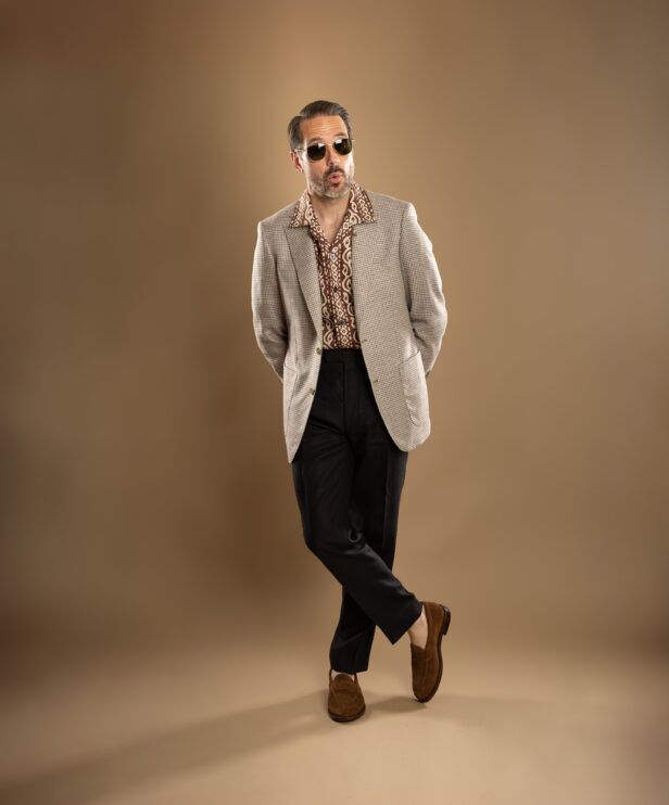 Beige houndstooth sport coat with brown printed camp collar shirt, black trousers and suede loafers, worn by a model