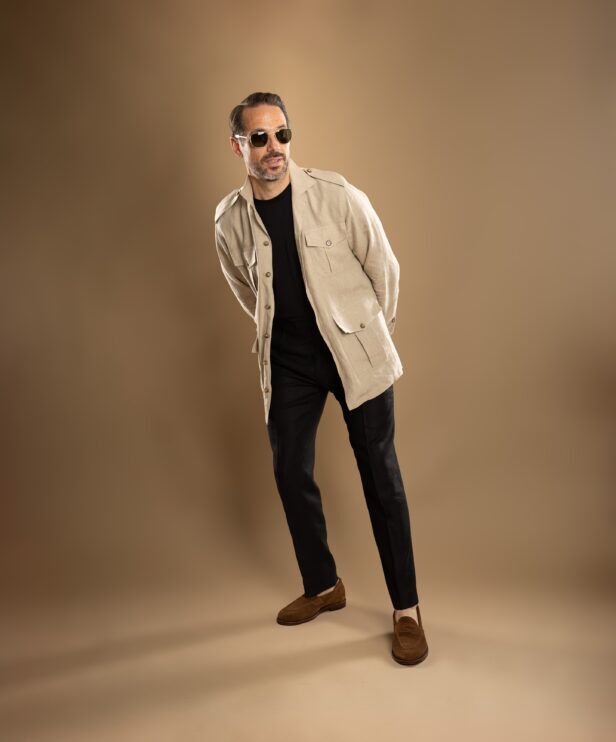 Beige linen safari jacket with patch pockets, worn open over a black tee, black trousers and brown suede loafers by a model