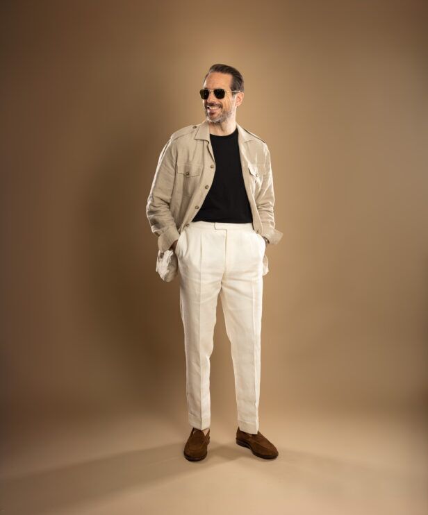 Beige linen safari jacket with black t-shirt, cream pleated trousers and brown suede loafers, worn by a model