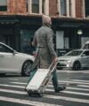 Grey Prince of Wales check overcoat and beanie, worn by a man pulling a silver suitcase across a crosswalk