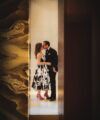 Black tuxedo with a bow tie beside a woman in a printed dress, kissing in a doorway