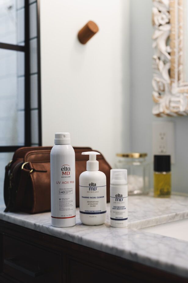 Three skincare bottles on a marble vanity beside a brown leather wash bag