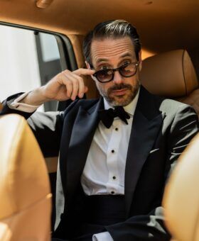 Black tuxedo with bow tie and stud front shirt, worn by a seated model in a car