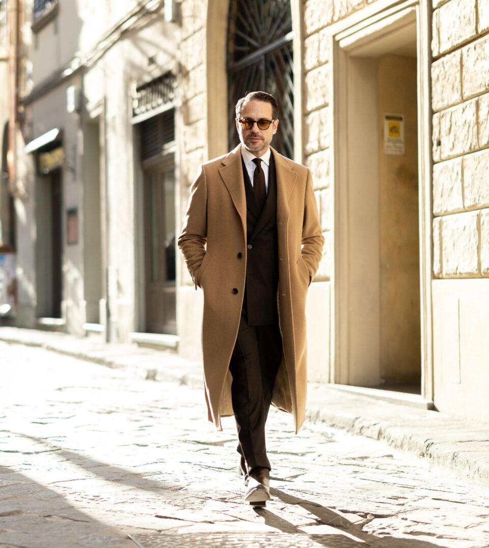 Camel overcoat over a dark brown double-breasted suit and knit tie, on a cobbled street