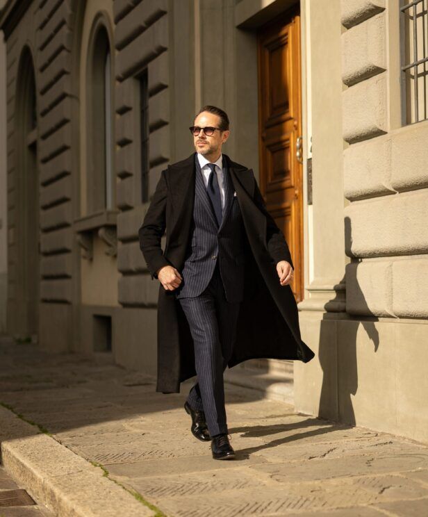 Black overcoat over a navy chalk stripe suit with navy tie, worn by a model walking