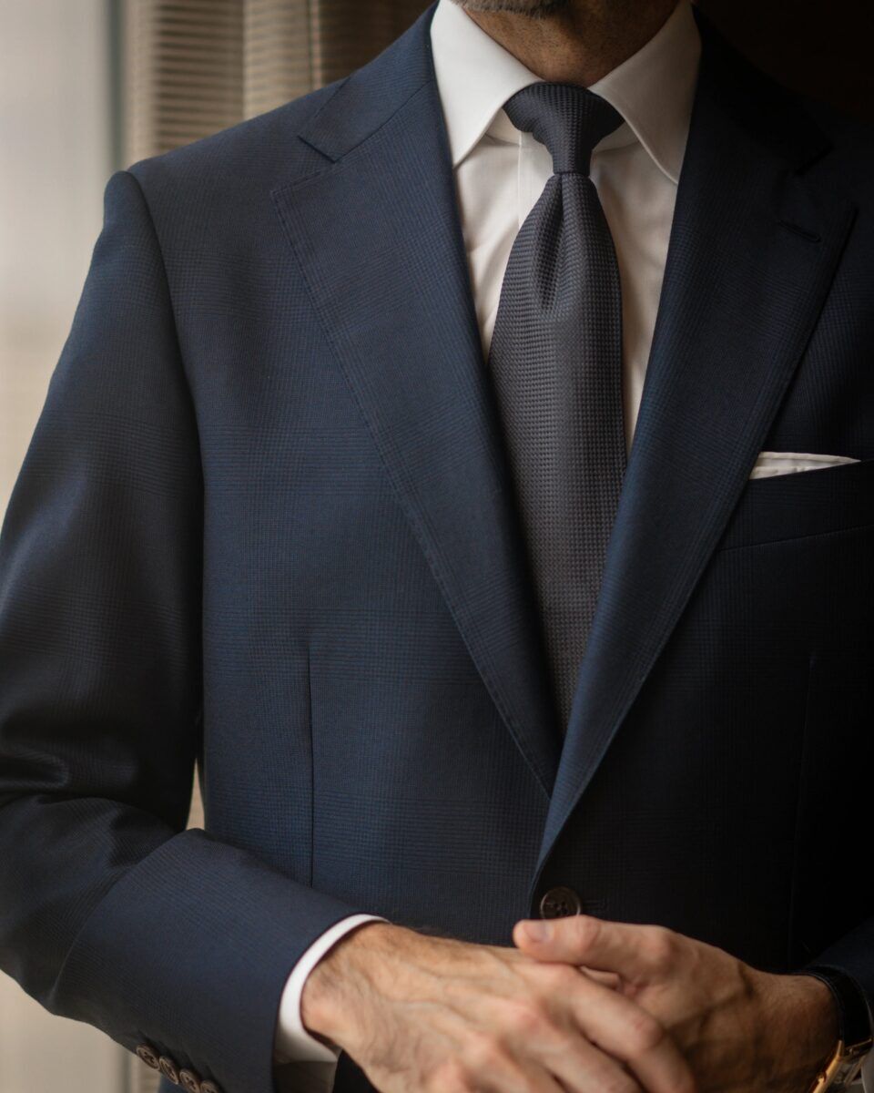 Navy subtle plaid wool suit jacket with peak lapel, white shirt and charcoal grenadine tie, worn by a model