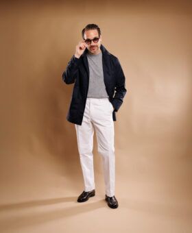 Navy twill chore coat with grey sweater, white trousers and brown loafers, full length