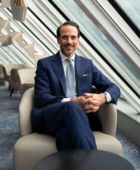 Royal blue birdseye suit with white shirt and pale blue knit tie, worn by a seated model