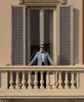 Light blue linen dinner jacket with navy bow tie, worn by a model on a shuttered balcony