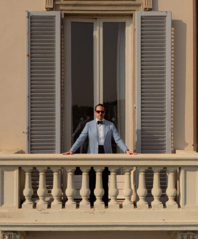 Light blue linen dinner jacket with navy bow tie, worn by a model on a shuttered balcony