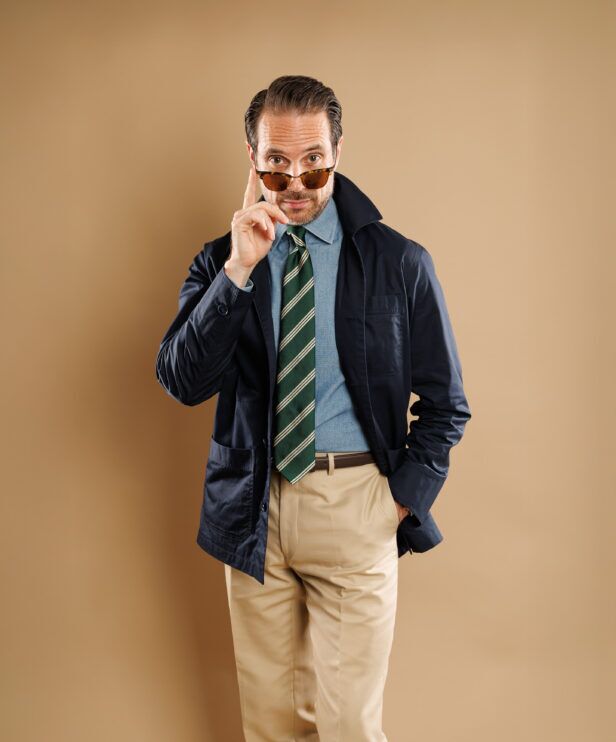 Navy chore coat over a chambray shirt with green striped tie and khaki trousers, worn by a model