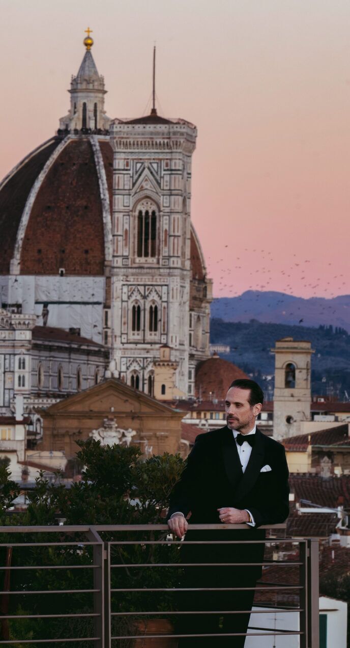 Black tuxedo with white shirt, black bow tie and pocket square, worn by a model on a Florence rooftop at dusk