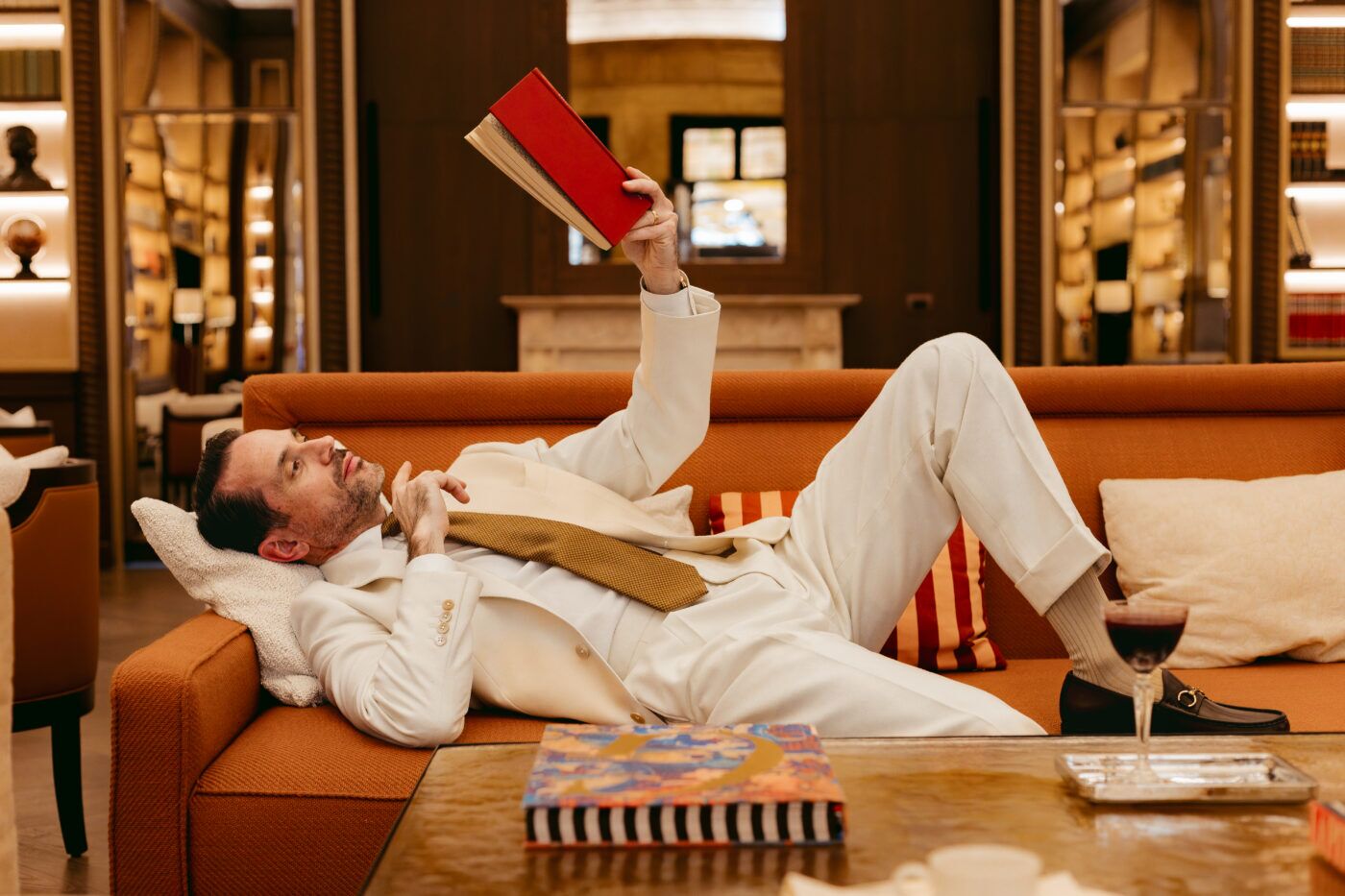 Cream double-breasted suit with gold knit tie and brown bit loafers, worn by a man reclining in a library lounge