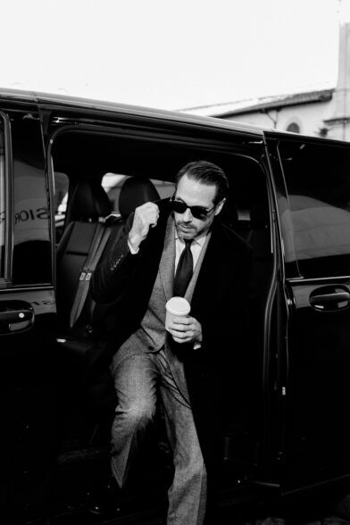 Glen check suit with dark overcoat, white shirt and knit tie, worn by a man stepping from a car in a black-and-white street scene