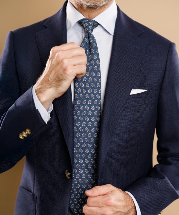 Blue silk necktie with tan and blue medallions, worn with navy hopsack blazer, pale blue shirt and white pocket square, by a model