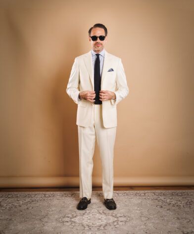 Ivory two-button suit with light blue shirt, navy knit tie and black horsebit loafers, worn by a model