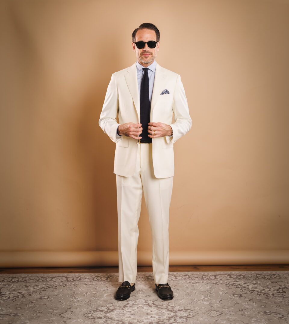 Ivory two-button suit with light blue shirt, navy knit tie and black horsebit loafers, worn by a model
