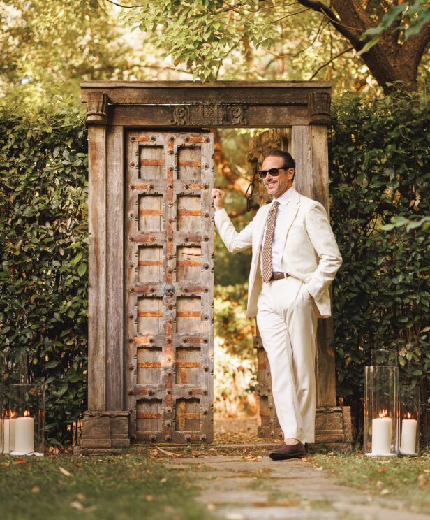 Ivory suit with white shirt, patterned tie and brown loafers, worn by a model by a garden gate