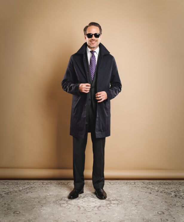 Navy raincoat over a charcoal suit with white shirt and purple dotted silk tie, worn by a model