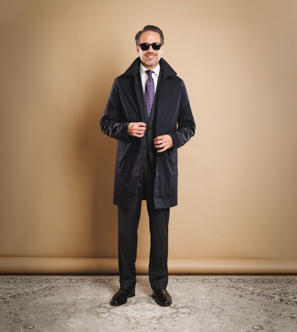 Navy raincoat over a charcoal suit with white shirt and purple dotted silk tie, worn by a model